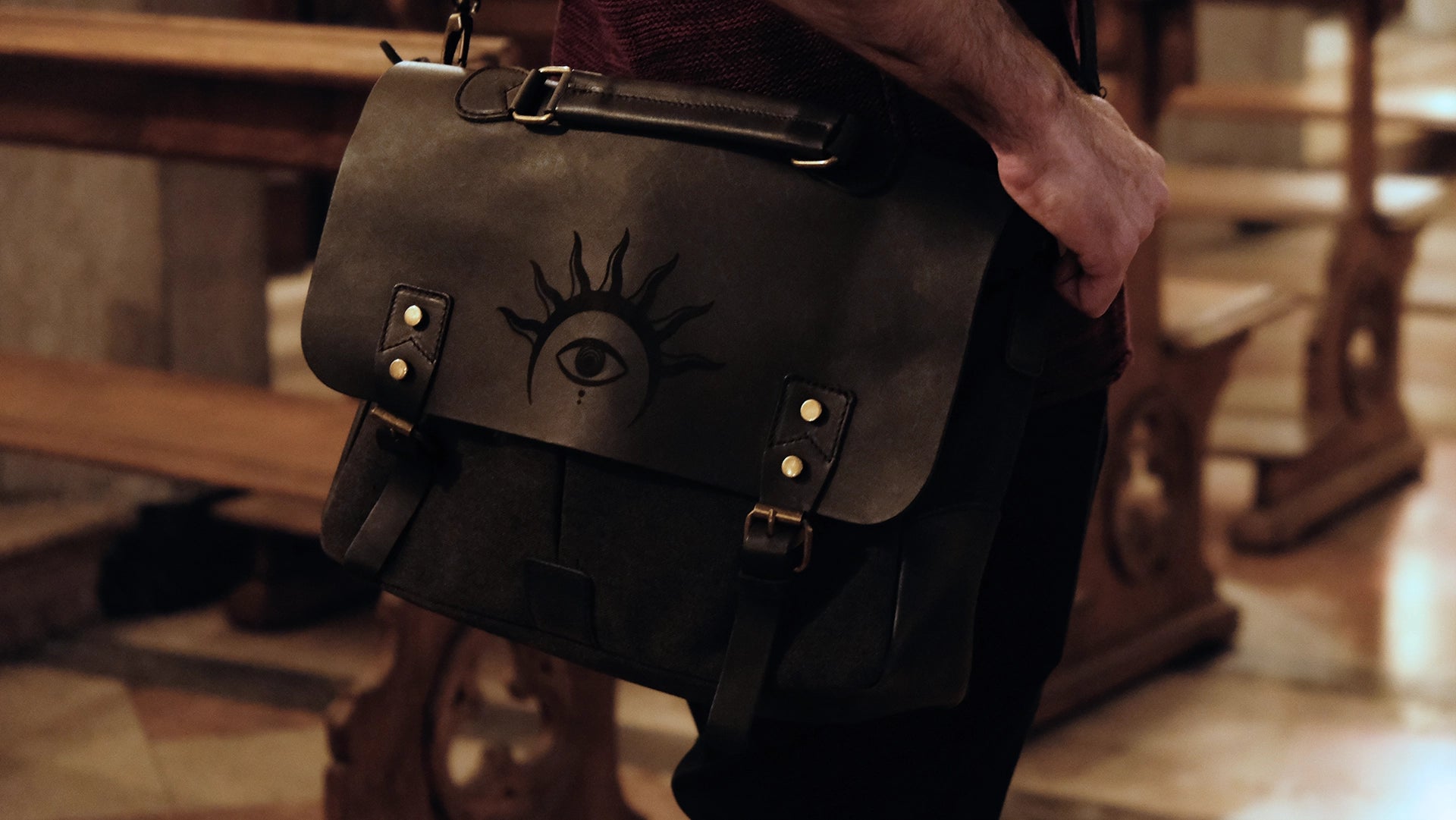 A model is wearing a black leather satchel engraved with a mystic eye with a solar crown. Dark academia style
