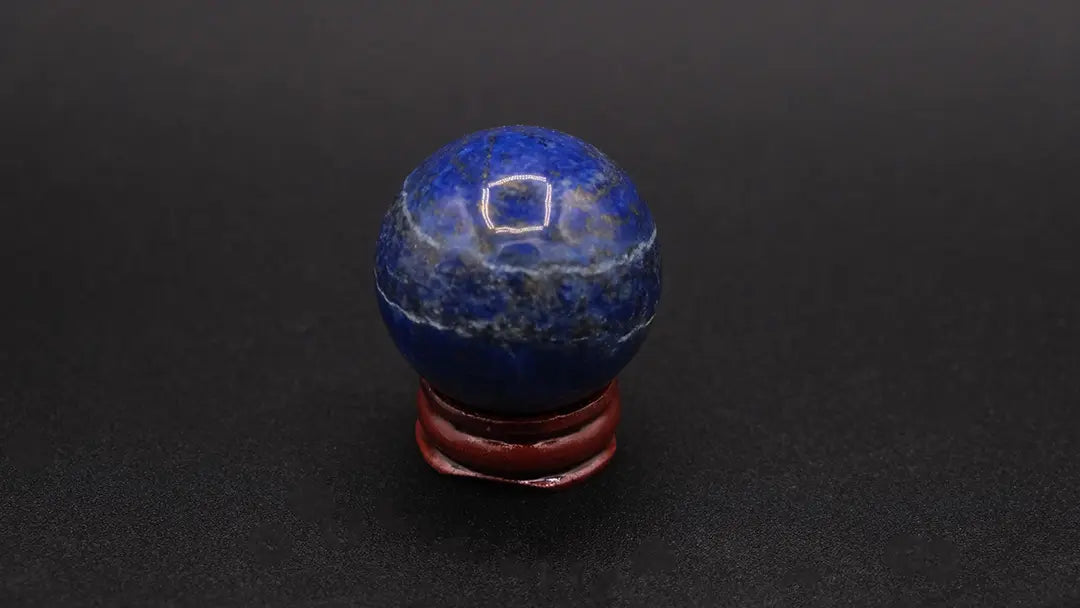 A perfect sphere made of blue lapis lazuli stone placed on a wooden stand over a black background