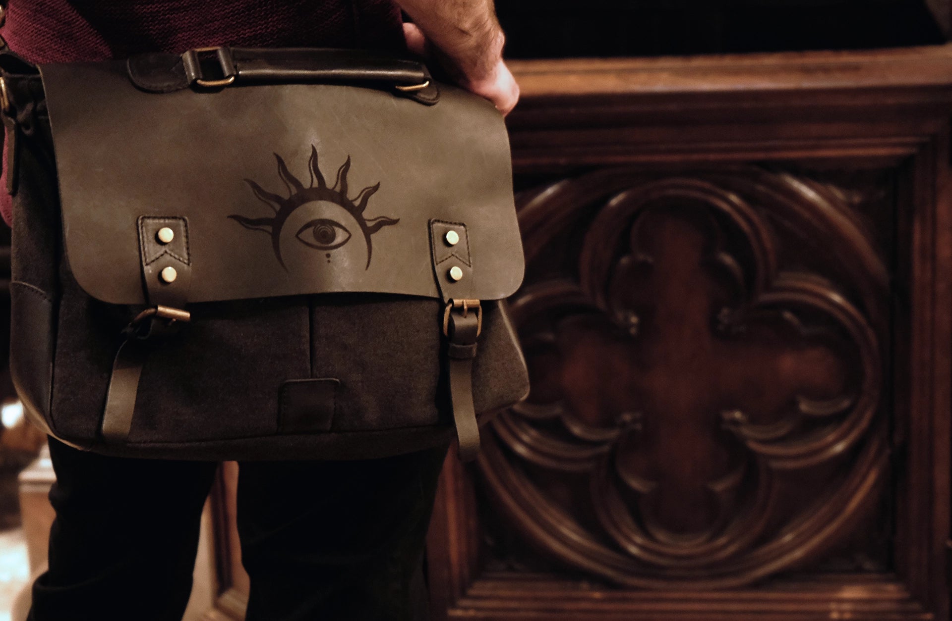 A model is wearing a black leather satchel engraved with a mystic eye with a solar crown. Dark academia style