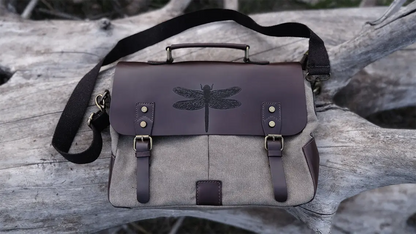 A brown leather satchel engraved with a dragonfly drawn is over a white tree