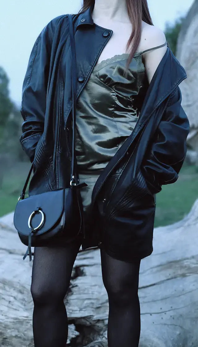 A girl wearing a green dress and a black leather jacket is posing with a black leather bag decorated with a golden ring and a slavic pattern